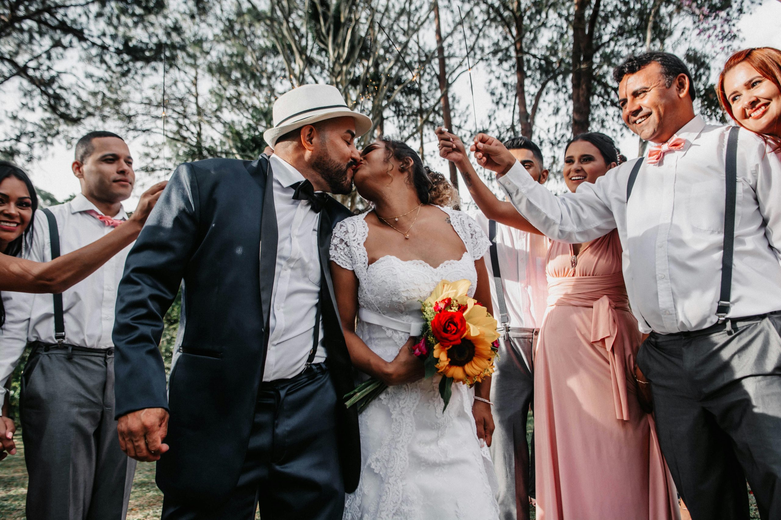 Newlywed couple kissing outdoors surrounded by joyful guests in wedding attire.